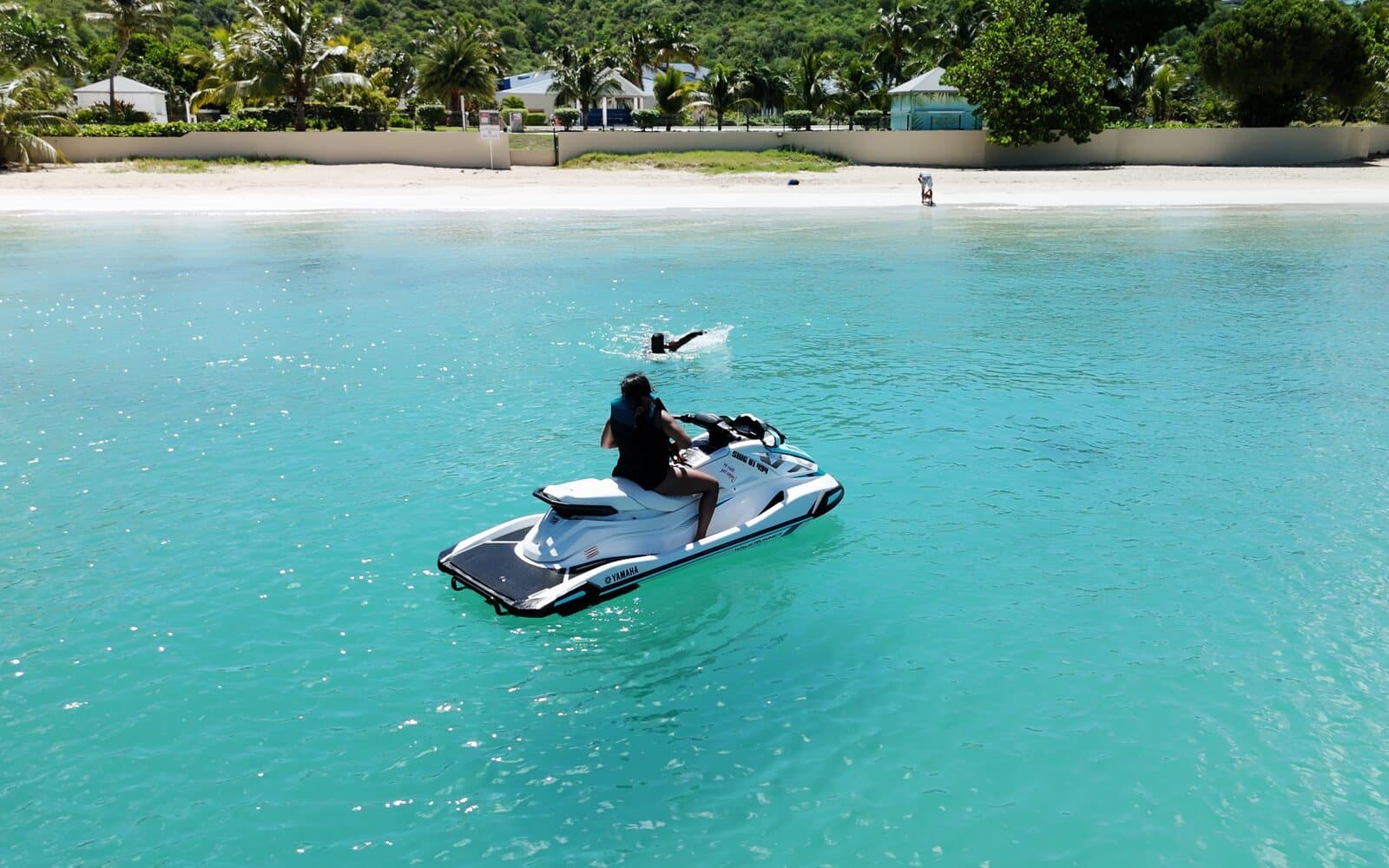 A passenger sitting on a stopped jet ski, a swimmer ahead of her, facing the beach