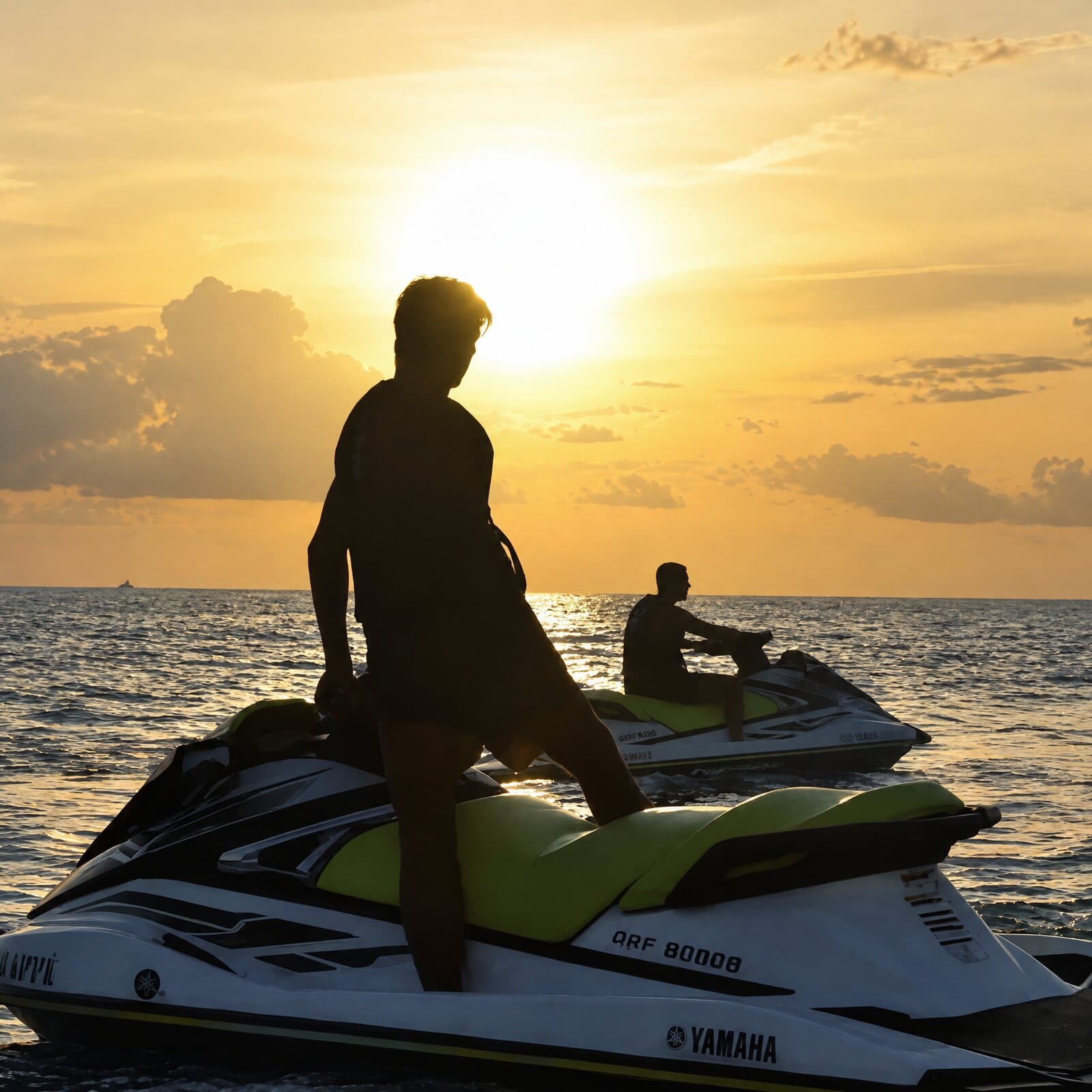 A rider standing on a jet ski, silhouetted against the setting sun, another jet ski behind him