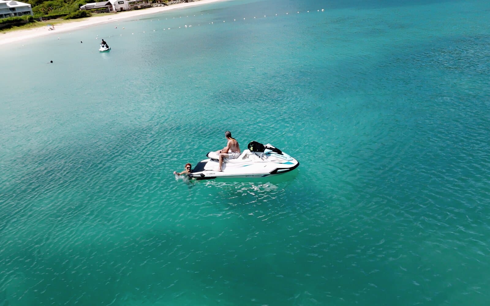 A man sitting on a stopped jet ski, a swimmer holding on at the back, seen from the drone
