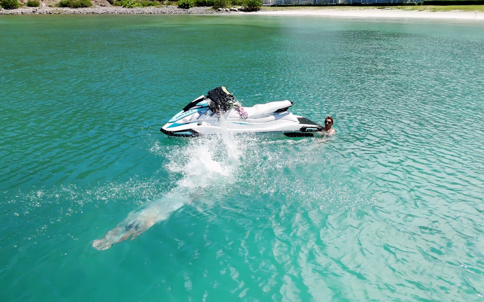A swimmer dives in next to a stopped jet ski on turquoise water, seen from the drone