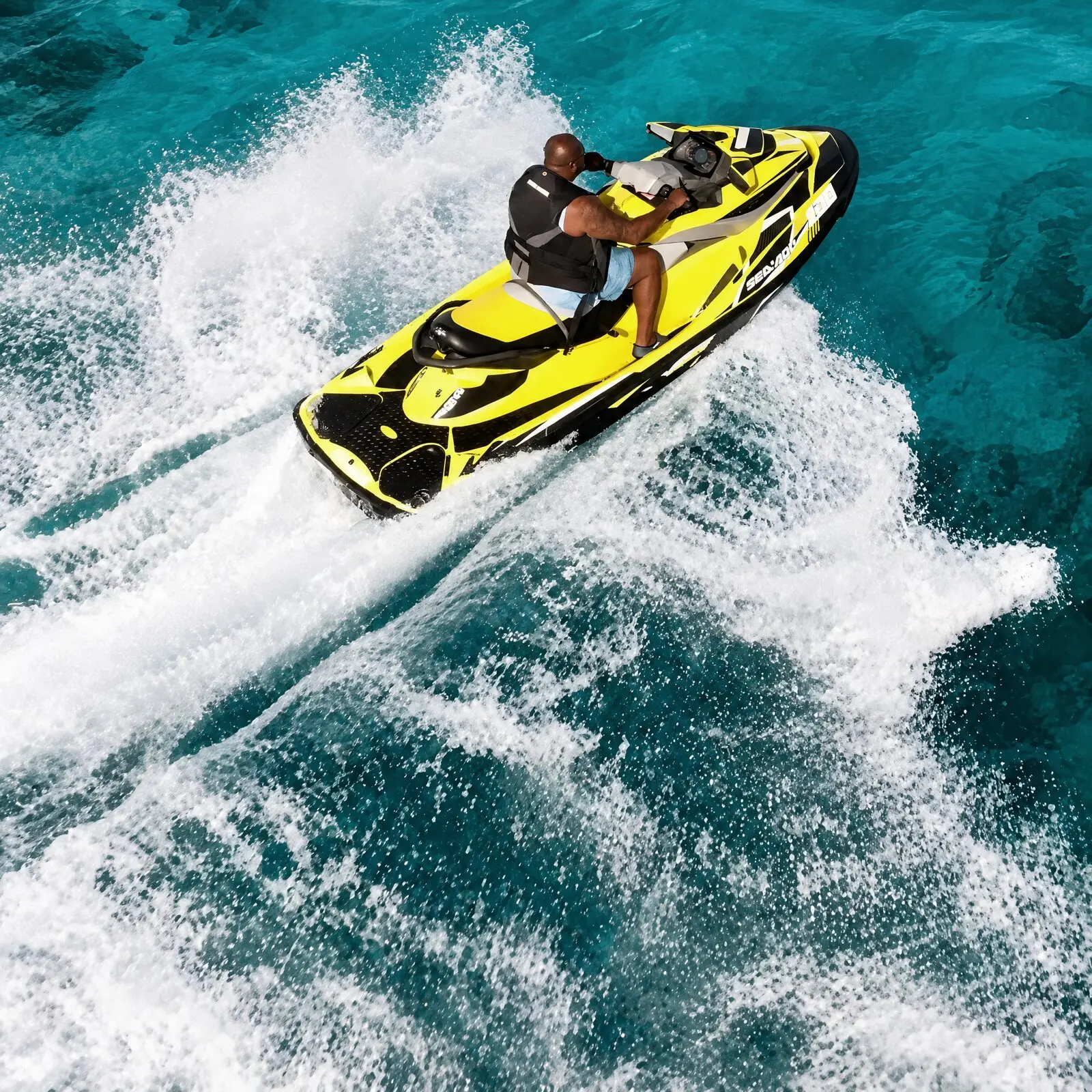 Dos personas en un jet ski amarillo que gira levantando una cortina de espuma, visto desde arriba