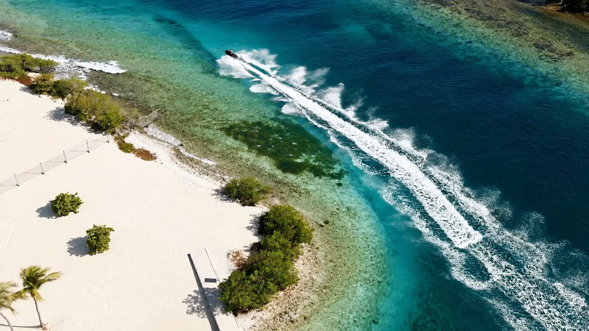 La estela de un jet ski bordea una playa de arena blanca, entre el agua turquesa y el azul del mar abierto, vista desde el cielo