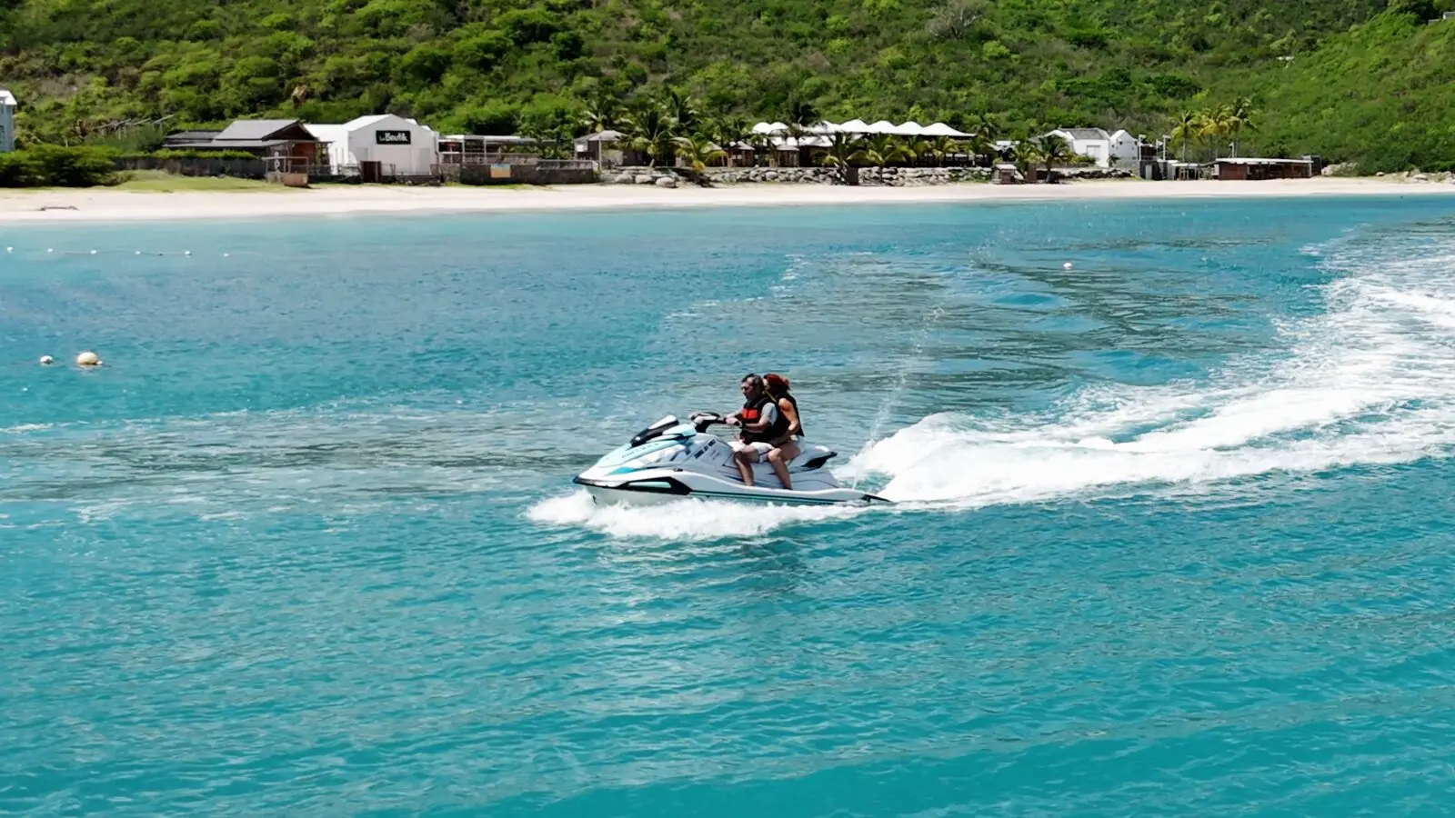 Deux personnes sur le même jet ski filent sur l'eau turquoise, une plage et des collines derrière