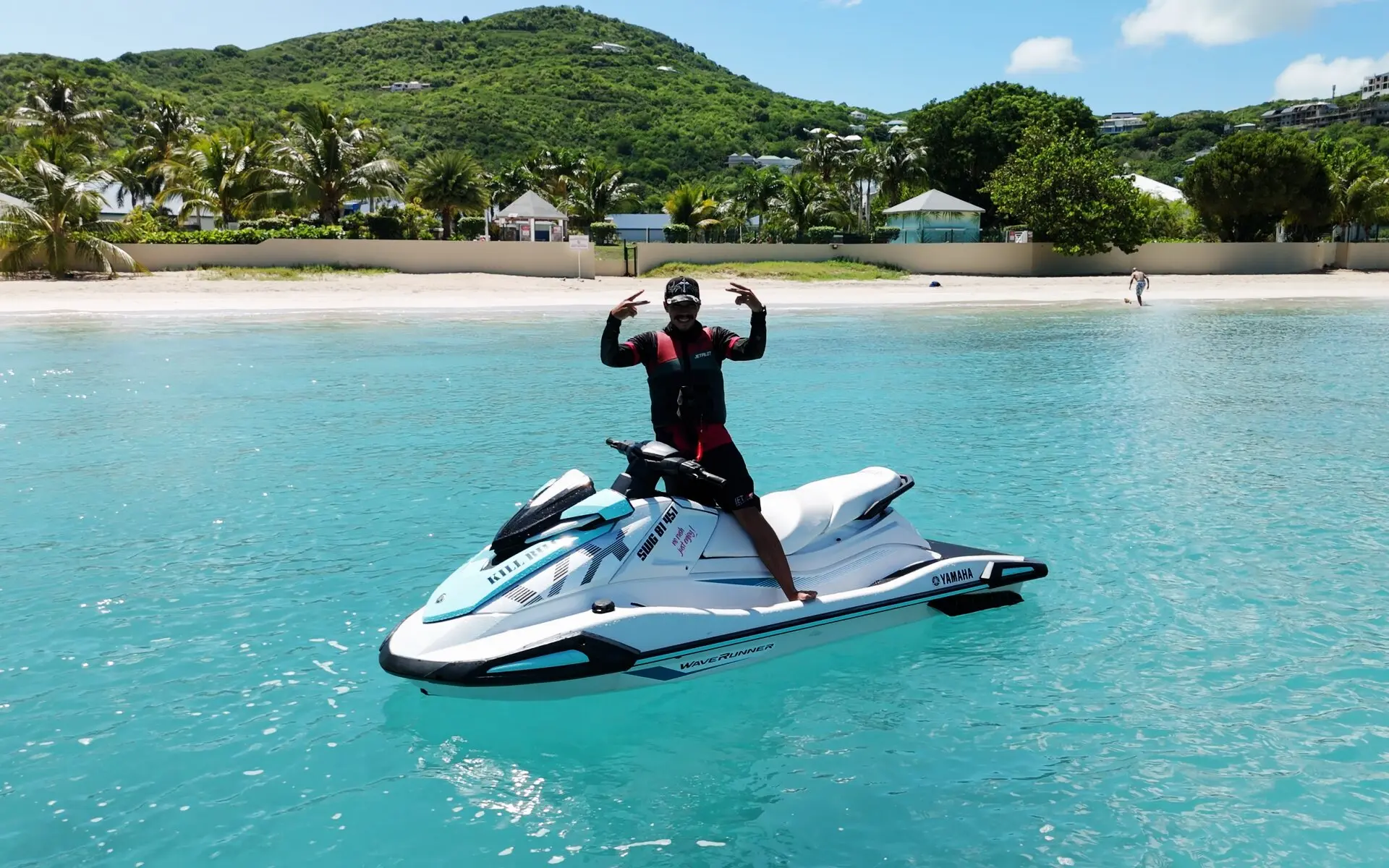 Un pilote debout sur son jet ski Yamaha blanc et turquoise, à l'arrêt dans l'eau claire devant une plage bordée de palmiers