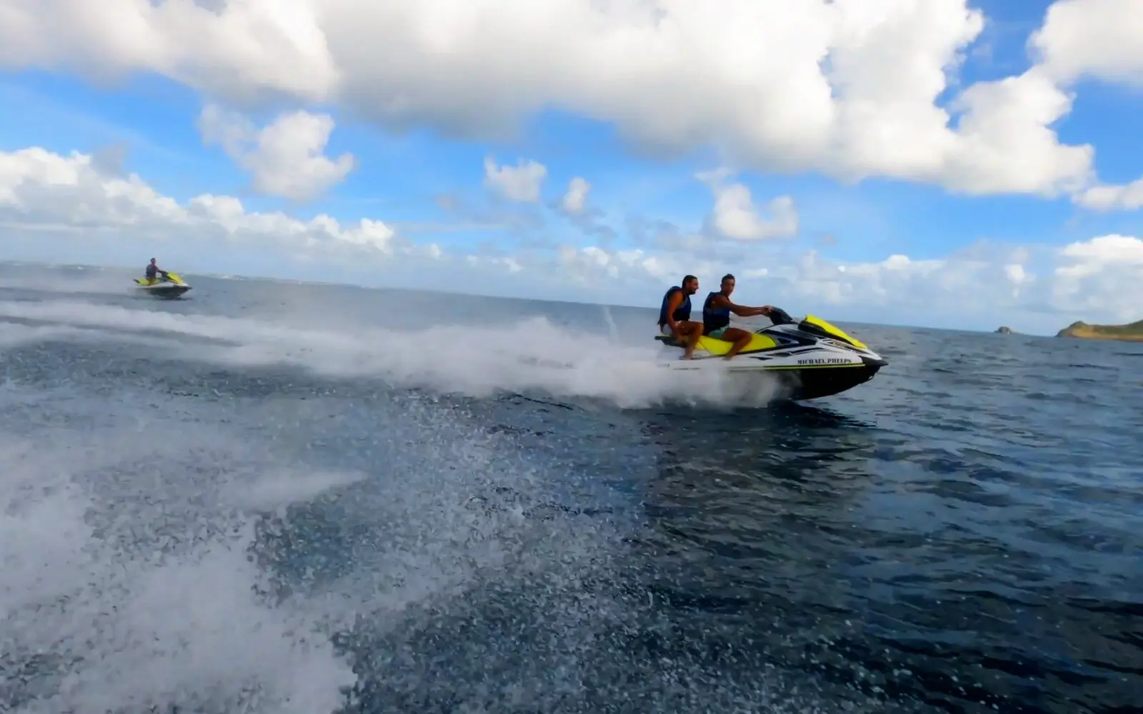 Dos personas en un jet ski a toda velocidad en mar abierto, con otro jet ski a lo lejos