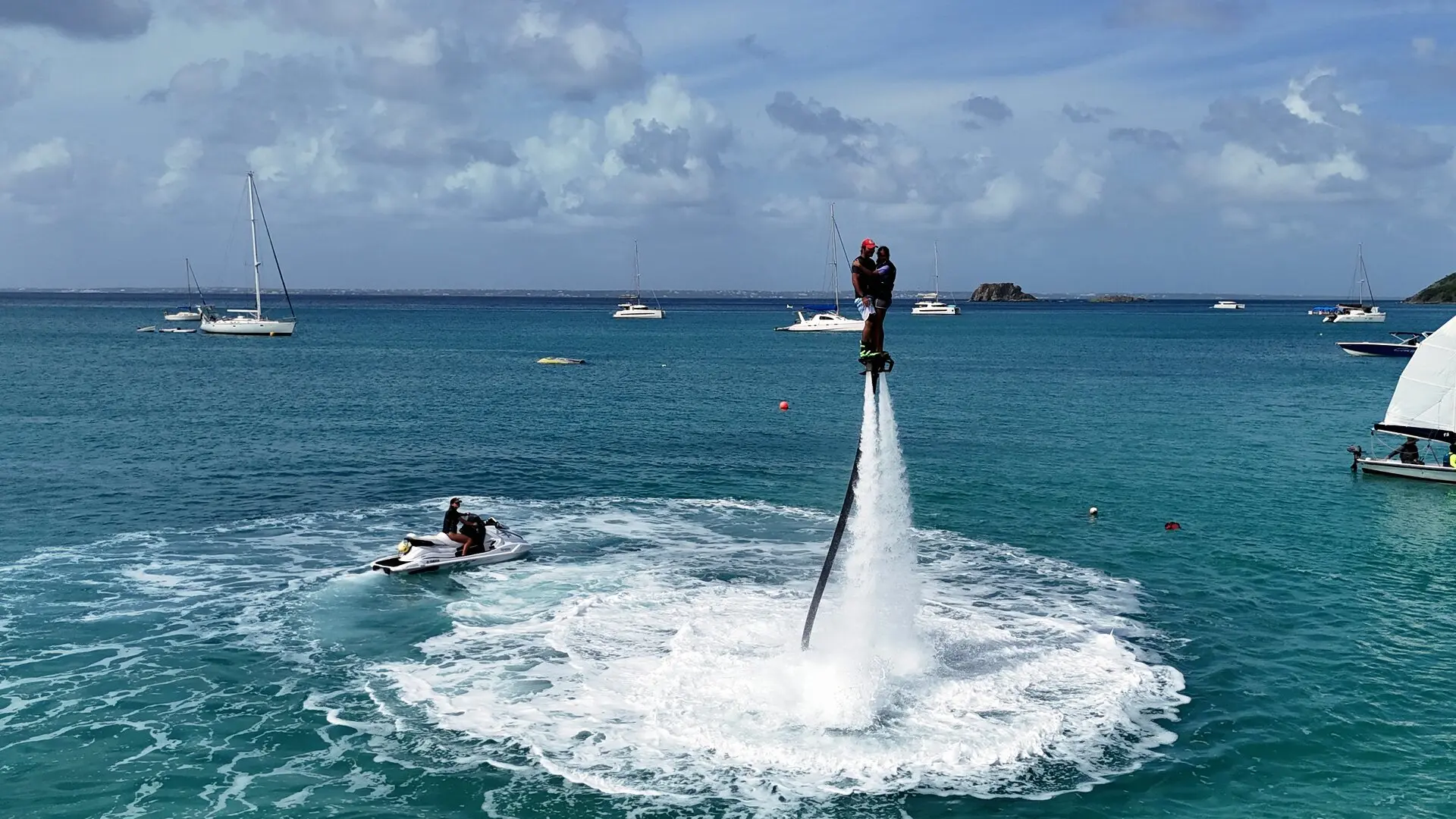 Dos personas en flyboard sobre el agua, un jet ski al lado y veleros fondeados