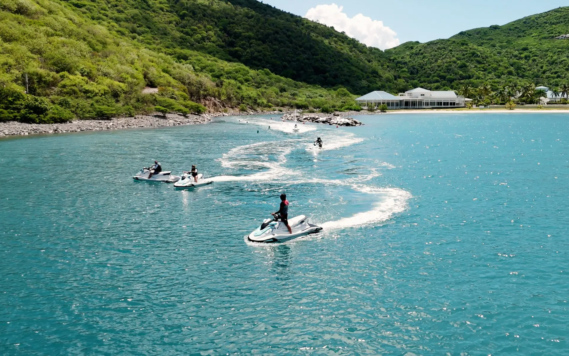 Des jet skis blancs à la file dans une baie turquoise, au pied de collines vertes
