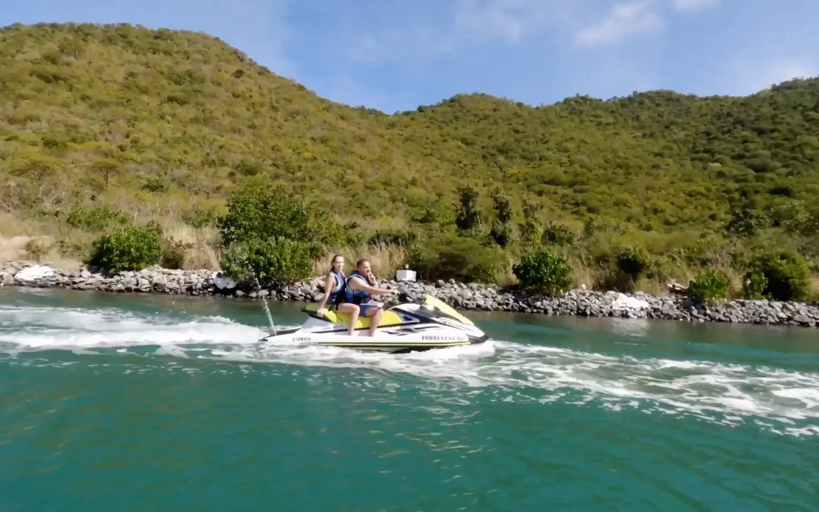 Dos personas en un jet ski amarillo, bordeando una costa verde