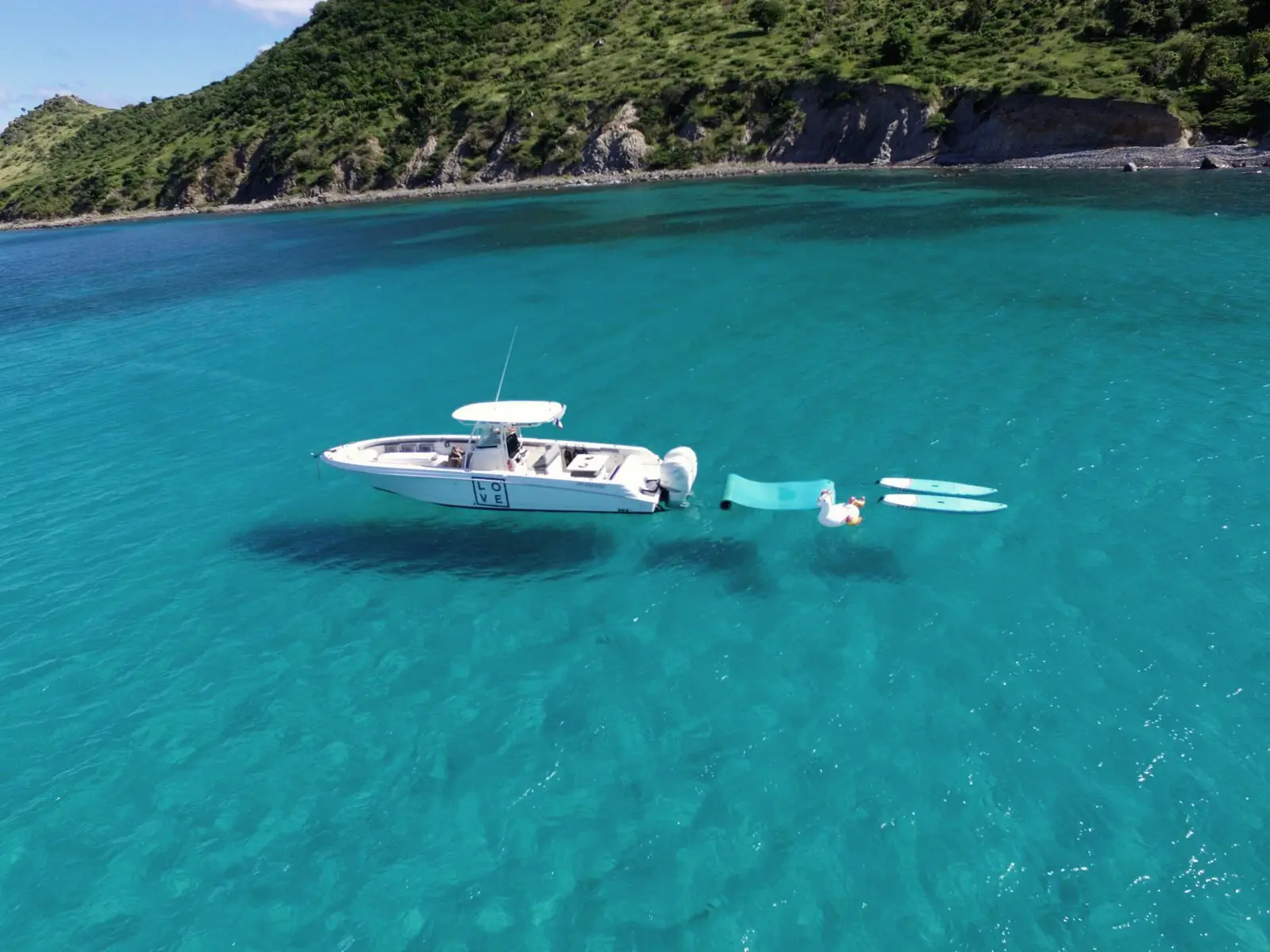 Un barco blanco fondeado en el agua turquesa, con una colchoneta flotante y tablas de paddle, visto desde el cielo